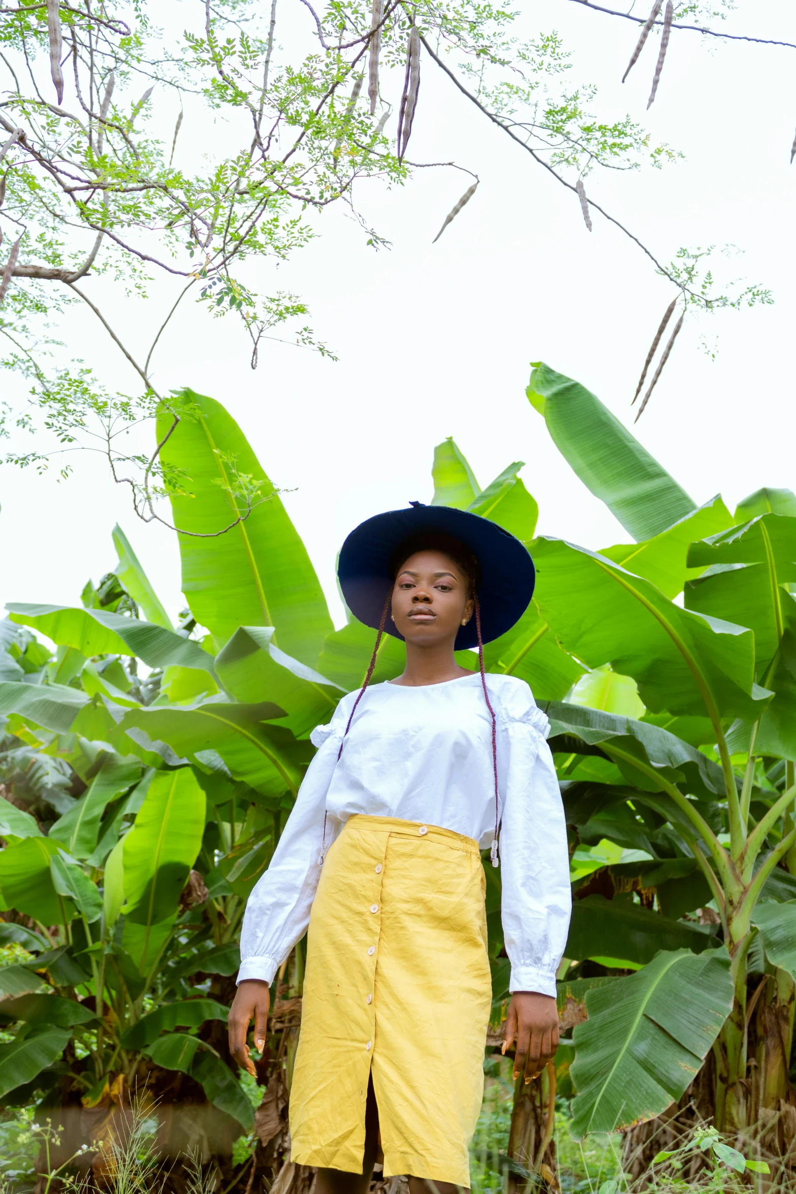 a woman standing in front of a banana tree, unsplash, afrofuturism, white shirt and green skirt, with hat, with yellow cloths, lush chic garden