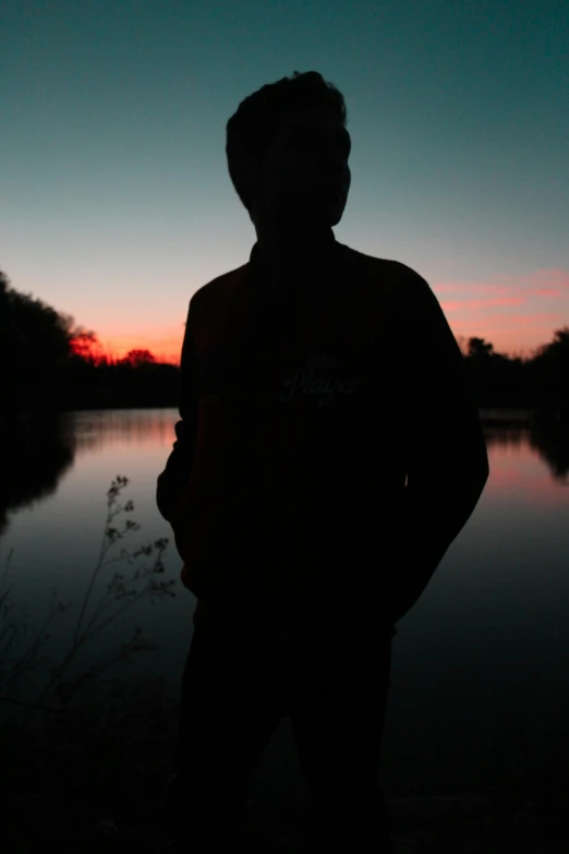 a man standing in front of a body of water, ((sunset)), around 1 9 years old, silhouette :7, andres rios