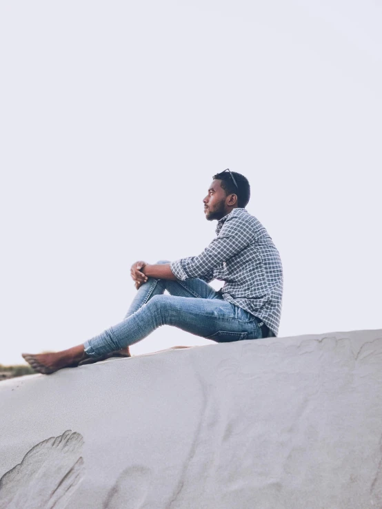 a man sitting on top of a sand dune, trending on unsplash, thoughtful pose, wearing a shirt and a jean, dark skinned, slightly pixelated