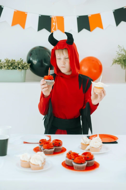 a woman in a devil costume sitting at a table with cupcakes, by Julia Pishtar, pexels, teen boy, on a white table, gif, kids