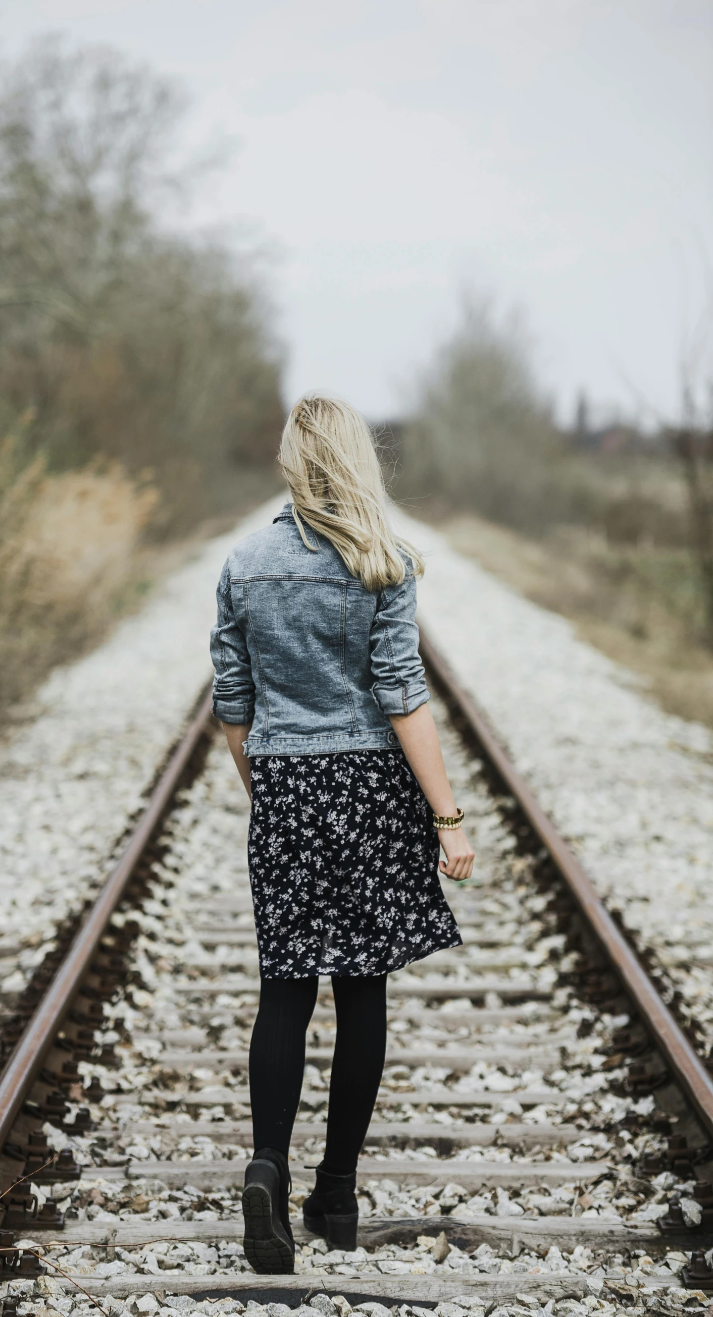 a woman is walking down a train track, pexels, jean jacket, patterned clothing, single file, grayish