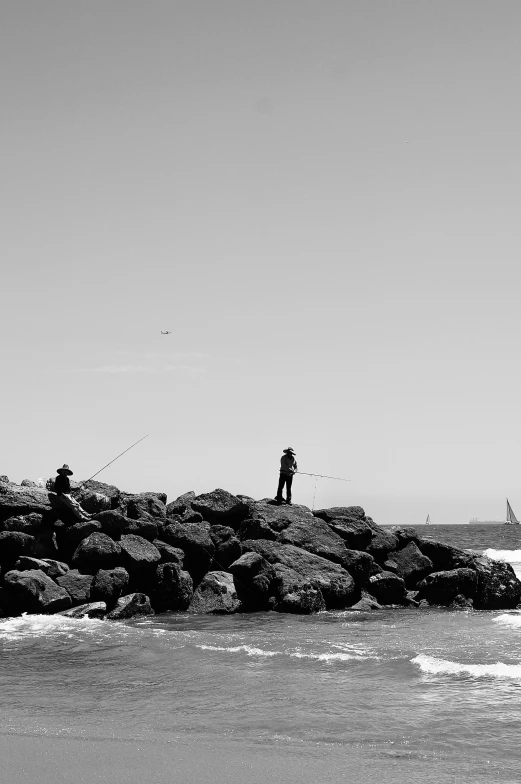 a man standing on top of a rock next to the ocean, a black and white photo, figuration libre, people angling at the edge, on a hot australian day, rocks flying, near a jetty