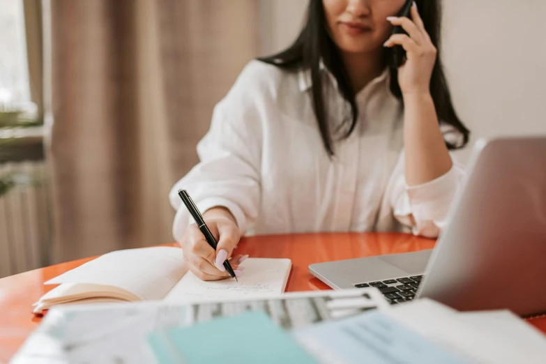 a woman sits at a table with an open notebook and pen, next to a laptop and cellphone, while holding a phone