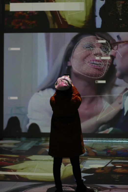 a person standing in front of a large screen, a hologram, inspired by Anna Füssli, interactive art, embracing, the mask covers her entire face, as she looks up at the ceiling, reuniting