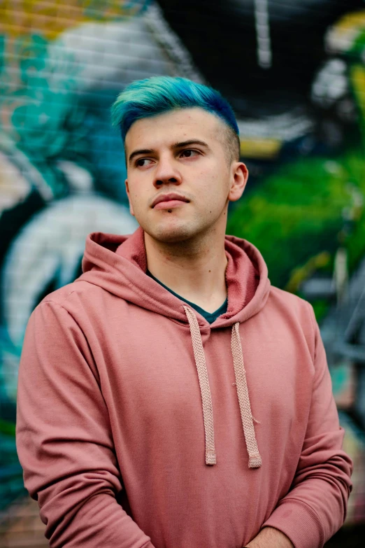 a man with blue hair standing in front of a graffiti wall, an album cover, by Jacob Toorenvliet, trending on pexels, wearing a pastel pink hoodie, headshot profile picture, luka mivsek, twitch streamer
