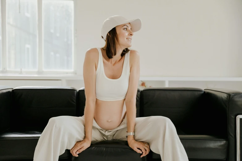 a woman sitting on top of a black couch, by Emma Andijewska, trending on pexels, happening, pregnant belly, white baseball cap, wearing a tracksuit, wearing a crop top