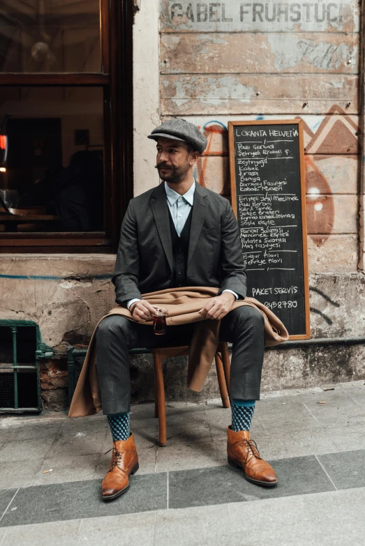 a man sitting on a chair in front of a building, dapper, sienna, in town, guide