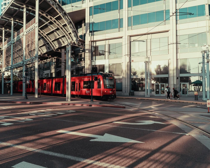 a red train traveling past a tall building, pexels contest winner, market street, street tram, thumbnail, afternoon light