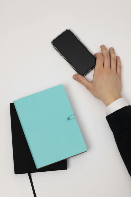 a person holding a cell phone next to a notebook, light-blue, no - text no - logo, hovering indecision, product shot