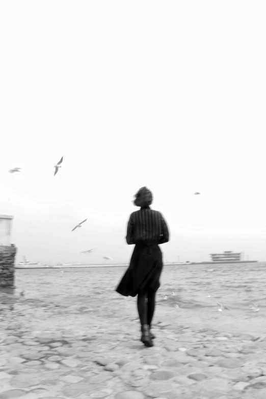 a black and white photo of a woman walking on the beach, inspired by Sergio Larraín, surrealism, flying birds in distance, **cinematic, black sea, harbor