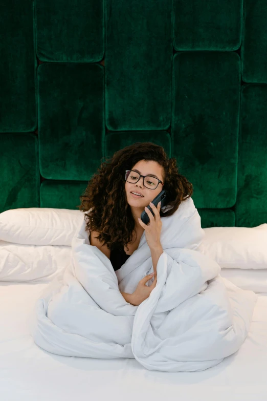 a woman sitting on a bed talking on a cell phone, curled up under the covers, wavy long black hair and glasses, 2019 trending photo, hotel room