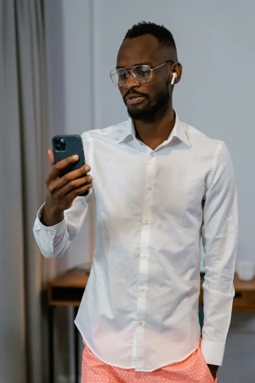 a man standing in front of a table holding a cell phone, trending on pexels, happening, wearing a white button up shirt, ( ( dark skin ) ), nerdy, wearing white pajamas