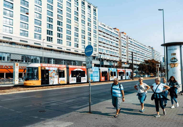 a group of people walking down a street next to a train, a photo, by Micha Klein, pexels contest winner, socialist realism, palast der republik in berlin, summer street near a beach, city panorama, 000 — википедия