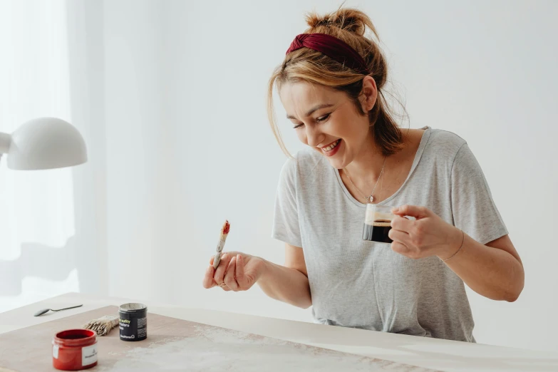 a woman sitting at a table painting on a piece of paper, trending on pexels, perfect smile vogue, celebration of coffee products, set against a white background, dark oil paint