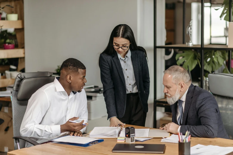 a group of people sitting around a wooden table, in an office