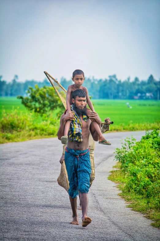 a man carrying a child on his back down a road, by Sudip Roy, wearing loincloth, bangladesh, road trip, carrying a saddle bag
