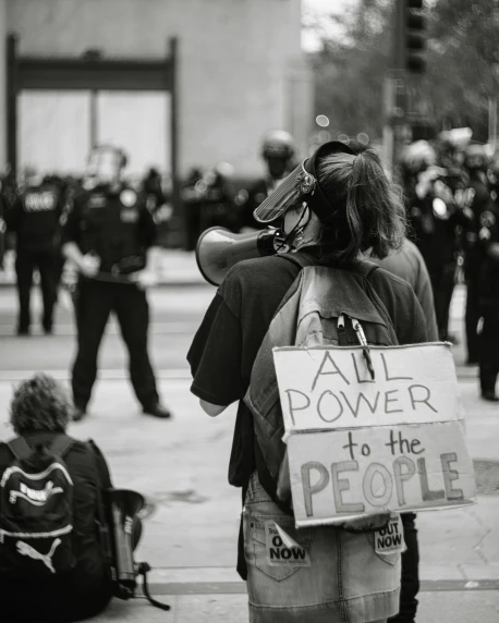 a woman holding a sign that says all power to the people, a black and white photo, trending on pexels, sots art, his back is turned, police, purpose is pump, a person standing in front of a