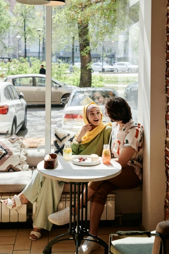 a couple of women sitting at a table in front of a window, by Basuki Abdullah, trending on pexels, people outside eating meals, hijab, scandinavian, sitting on a mocha-colored table
