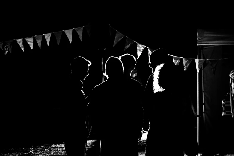 a group of people standing in front of a tent, a black and white photo, by Robin Guthrie, pexels, the lighting is dark, profile picture 1024px, silhouetted, dark people discussing