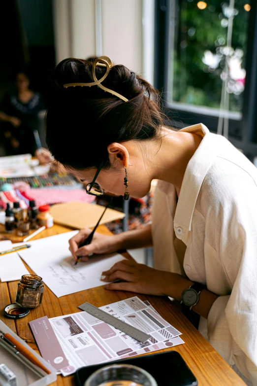 a woman sitting at a table writing on a piece of paper, process art, crafts and souvenirs, superfine inking, thumbnail, fashion designer