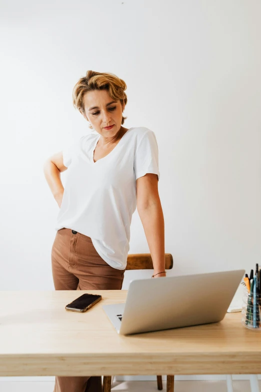 a woman standing in front of a laptop computer, by Nicolette Macnamara, trending on pexels, figuration libre, knees upturned, wearing a light shirt, in the office, concern