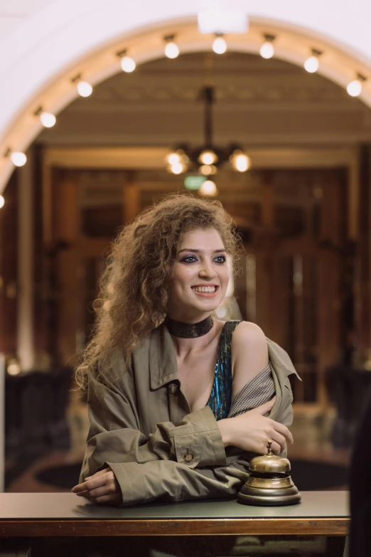 a woman that is sitting at a table, an album cover, brown curly hair, wearing a fancy jacket, ekaterina, while smiling for a photograph