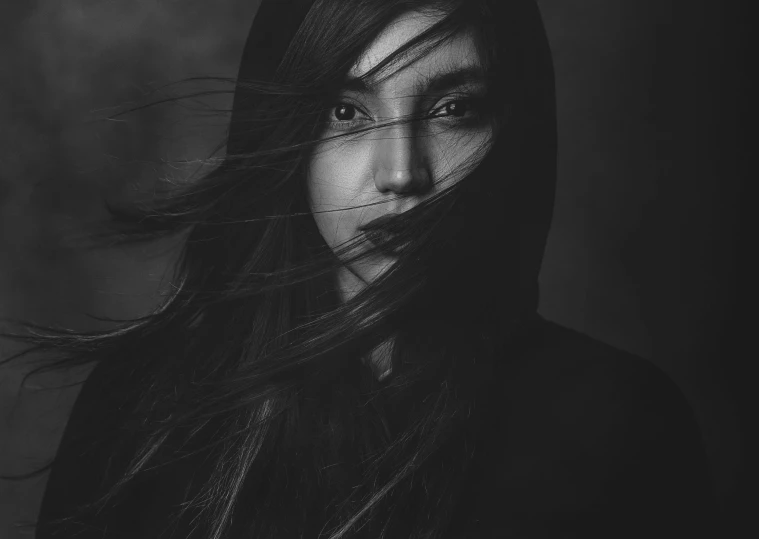 a woman with her hair blowing in the wind, a black and white photo, by Adam Marczyński, pexels contest winner, with haunted eyes and dark hair, asian woman, wearing a dark hood, studio potrait