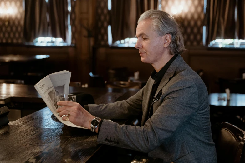 a man sitting at a table reading a newspaper, a portrait, by Emma Andijewska, unsplash, private press, henrik fisker, sitting at the bar, gray haired, elegantly dressed