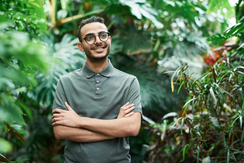 a man in glasses standing with his arms crossed by some trees