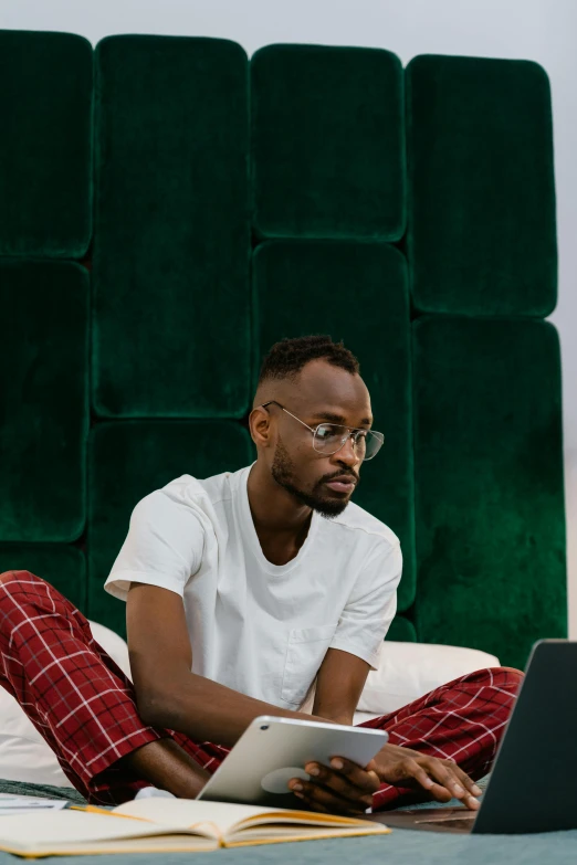 a man sitting on a bed using a laptop, by Cosmo Alexander, trending on pexels, african aaron paul, wearing small round glasses, red and green tones, contemplative