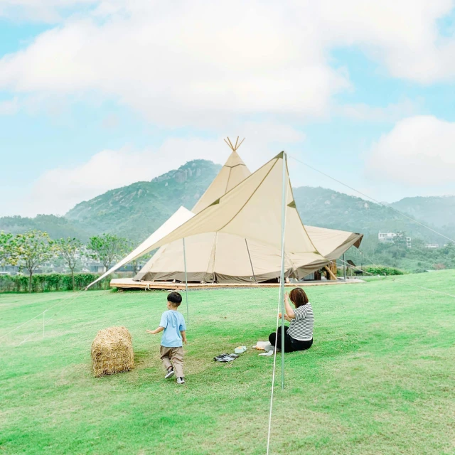 a couple of people sitting on top of a lush green field, gutai group, teepee, covered outdoor stage, jin shan, family friendly