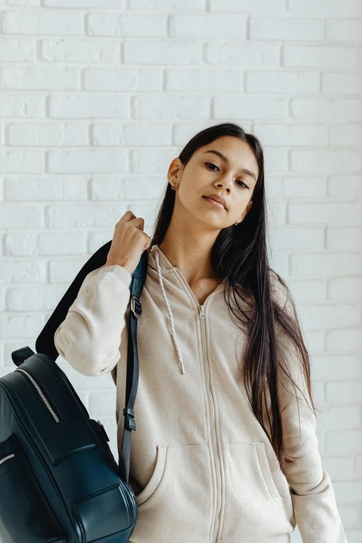 a woman standing in front of a white brick wall, trending on pexels, black haired girl wearing hoodie, with a backpack, young with long hair, wearing a light shirt