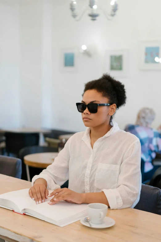 a woman sitting at a table reading a book, trending on unsplash, visual art, wearing shades, standing in class, looking serious, non binary model