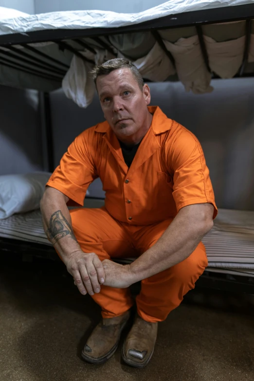 a man sitting on top of a bunk bed, a tattoo, wearing orange prison jumpsuit, col price, sean harris, 4k still