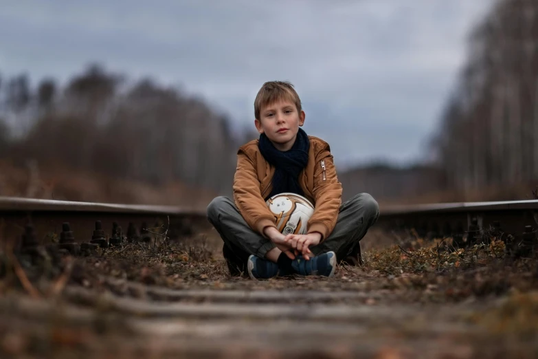 a boy in a brown jacket is crouching on the ground with a soccer ball