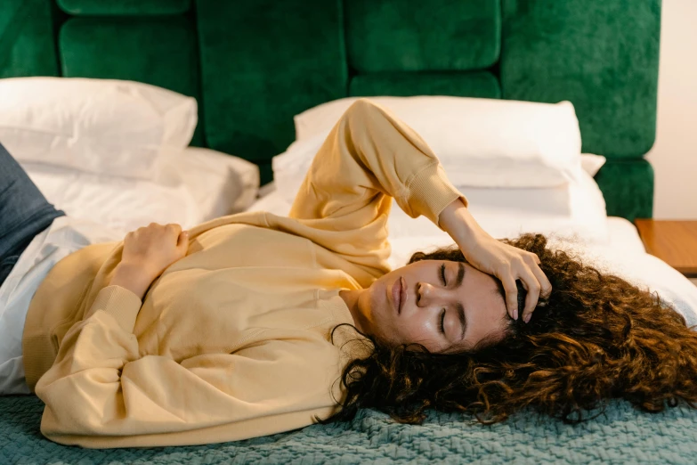 a woman laying on top of a bed next to a green headboard, by Julia Pishtar, trending on pexels, happening, yellow robe, frustrated detailed, brown curly hair, acupuncture treatment
