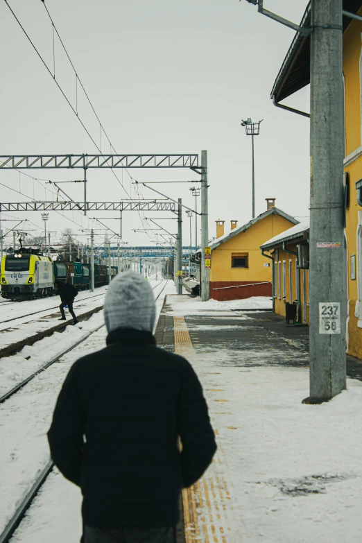 a person standing at a train station in the snow, by Anton Lehmden, small town surrounding, yellow, balaclava, back to camera