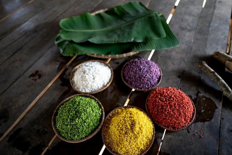 a wooden table topped with bowls filled with different colored rice, a portrait, inspired by Asai Chū, unsplash, hurufiyya, tropical leaves, purple sand, detailed product image, 1960s-era