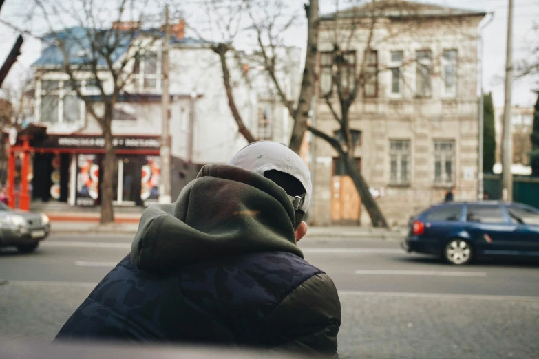 a man that is standing in the street, a photo, unsplash, realism, man sitting facing away, in a hood, cold sunny weather, building in the distance