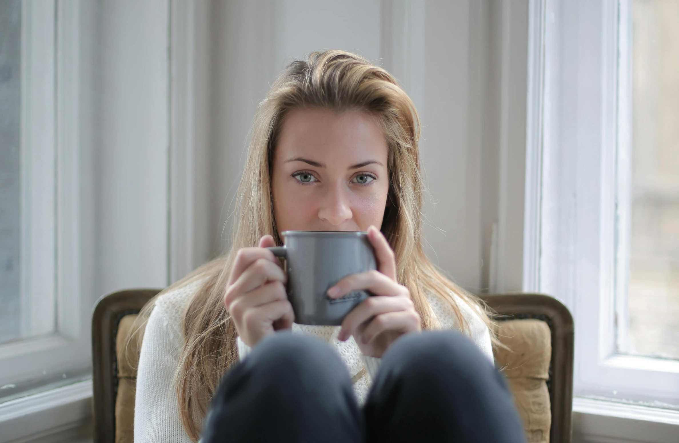 a woman sitting in a chair holding a cup, pexels, grey sweater, is ((drinking a cup of tea)), blonde, thoughtful