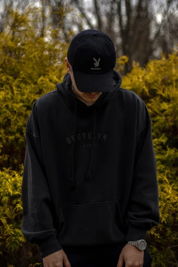 a man in a black hoodie standing in front of a bush, unsplash, stuckism, baggy clothing and hat, official store photo, faded hat, header with logo