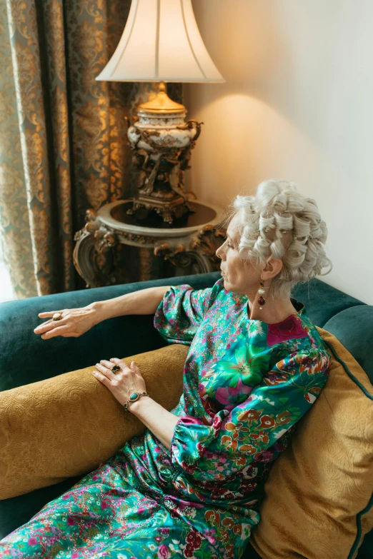 a woman sitting on a couch next to a lamp, by Joanna Carrington, trending on pexels, rococo, curled silver hair, patterned clothing, meditative sacral pose, armchairs