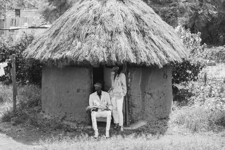 a black and white photo of two men in front of a hut, by Maurycy Gottlieb, pexels, visual art, african domme mistress, 1987 photograph, sitting, portrait of tall