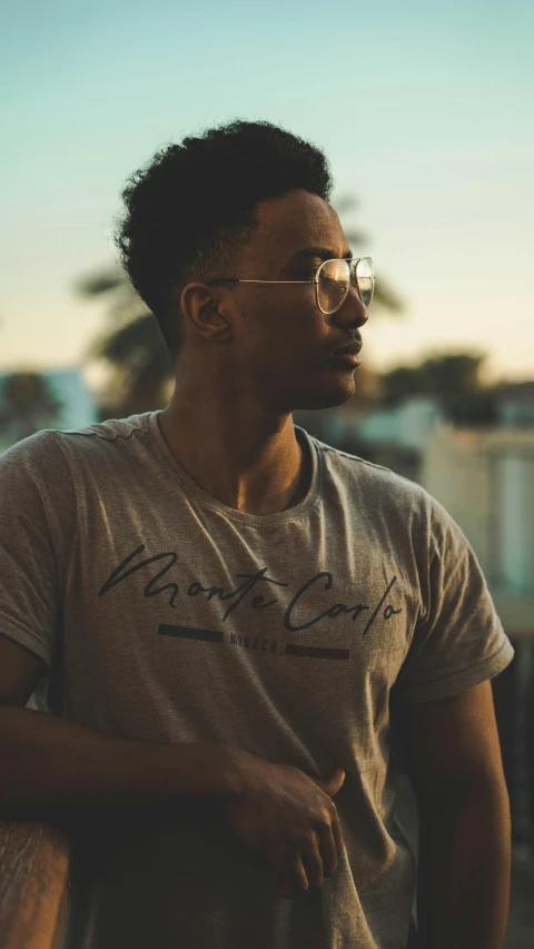 a man wearing glasses standing next to a railing, an album cover, by Cosmo Alexander, pexels contest winner, wearing a t-shirt, profile image, light skin, backlighted