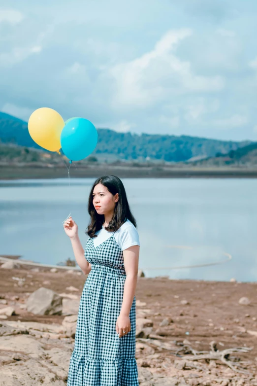 a woman standing on a beach holding a bunch of balloons, by Tan Ting-pho, pexels contest winner, near a lake, korean girl, icon, blue and yellow