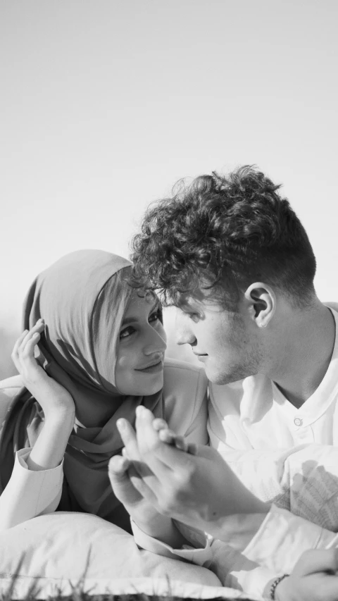 a man and woman sitting on a bed looking at a cell phone, a black and white photo, by Ismail Acar, pexels, hurufiyya, white hijab, handsome face and beautiful face, on beach, ( redhead