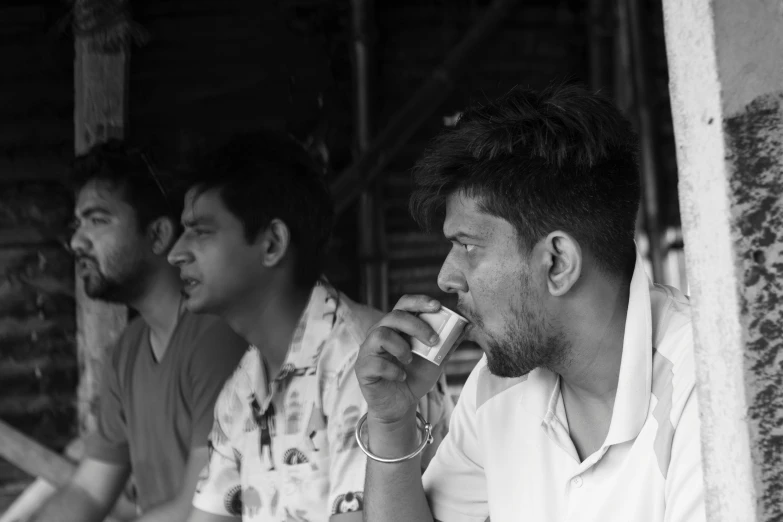 a group of men sitting next to each other, a black and white photo, by Jitish Kallat, pexels, the straw is in his mouth, holding a drink, left profile, shady look