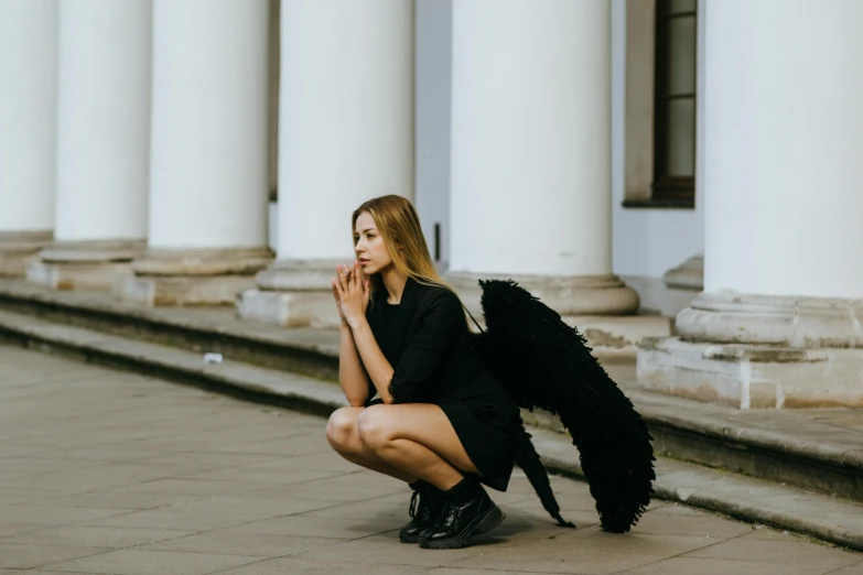 a woman kneeling on the ground in front of a building, by Emma Andijewska, pexels contest winner, magic realism, villainess has black angel wings, cara delevingne standing, slightly minimal, halloween