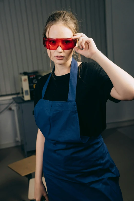 a woman in a blue apron holding a pair of red glasses, by Adam Marczyński, pexels contest winner, arbeitsrat für kunst, in an underground laboratory, streetwear, red laser, plating