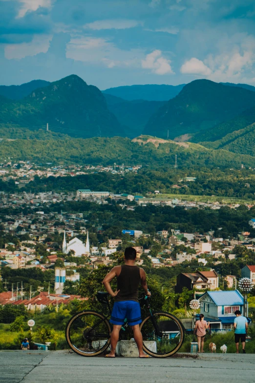 a man standing next to a bike on top of a hill, sumatraism, mountains and a huge old city, overlooking, blue, lush surroundings